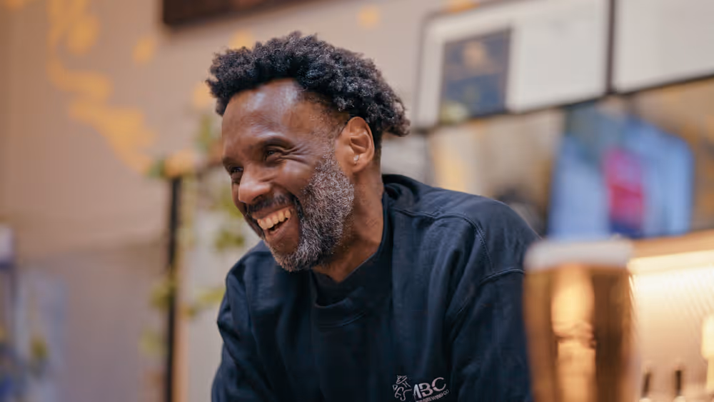 Smiling man with curly hair and gray beard wearing a dark shirt in a cozy, decorated room.