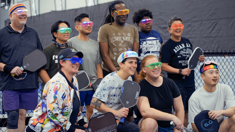 Group of pickleball players smiling, holding paddles, wearing colorful shutter shades and headbands indoors.