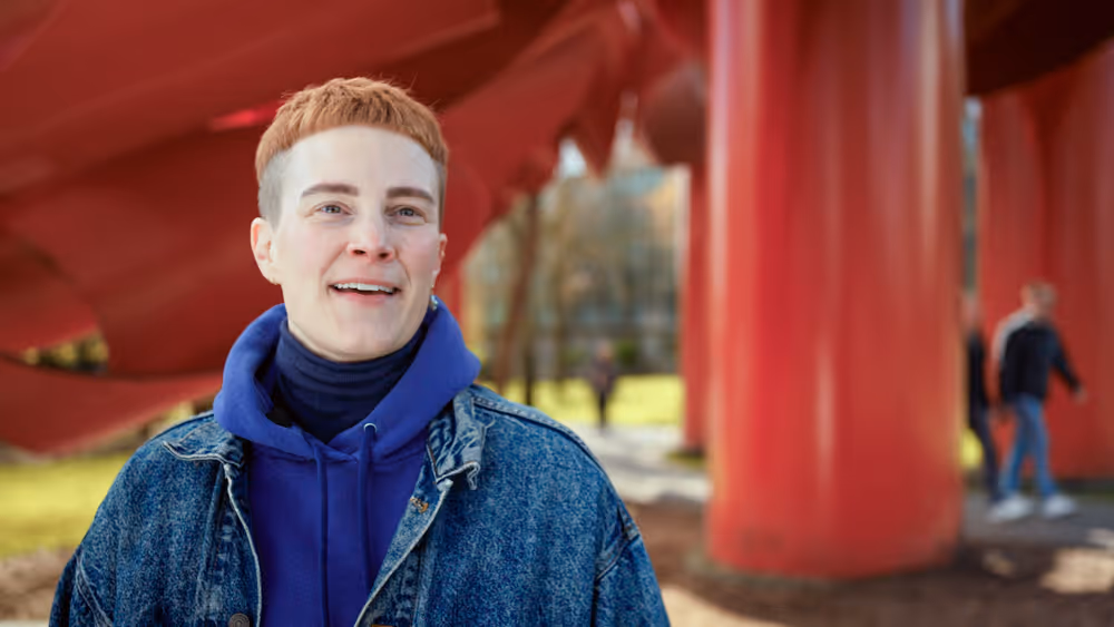 Person with short red hair wearing denim jacket and blue hoodie outdoors near large red sculpture.
