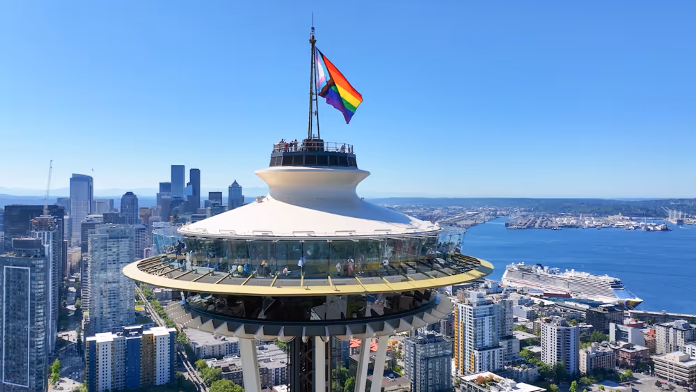 Seattle Space Needle with a flying Pride rainbow flag against a clear blue sky and cityscape background.