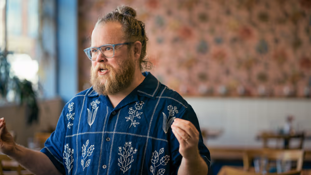 Man with beard and glasses speaking with expressive hand gestures in a brightly lit room.