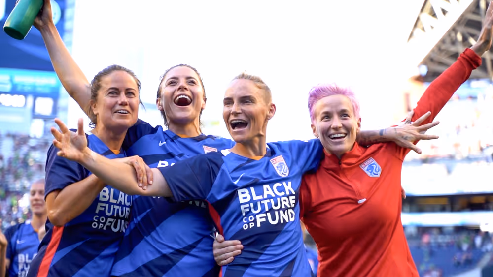 Four female soccer players smiling and celebrating together on the field in blue and red uniforms.