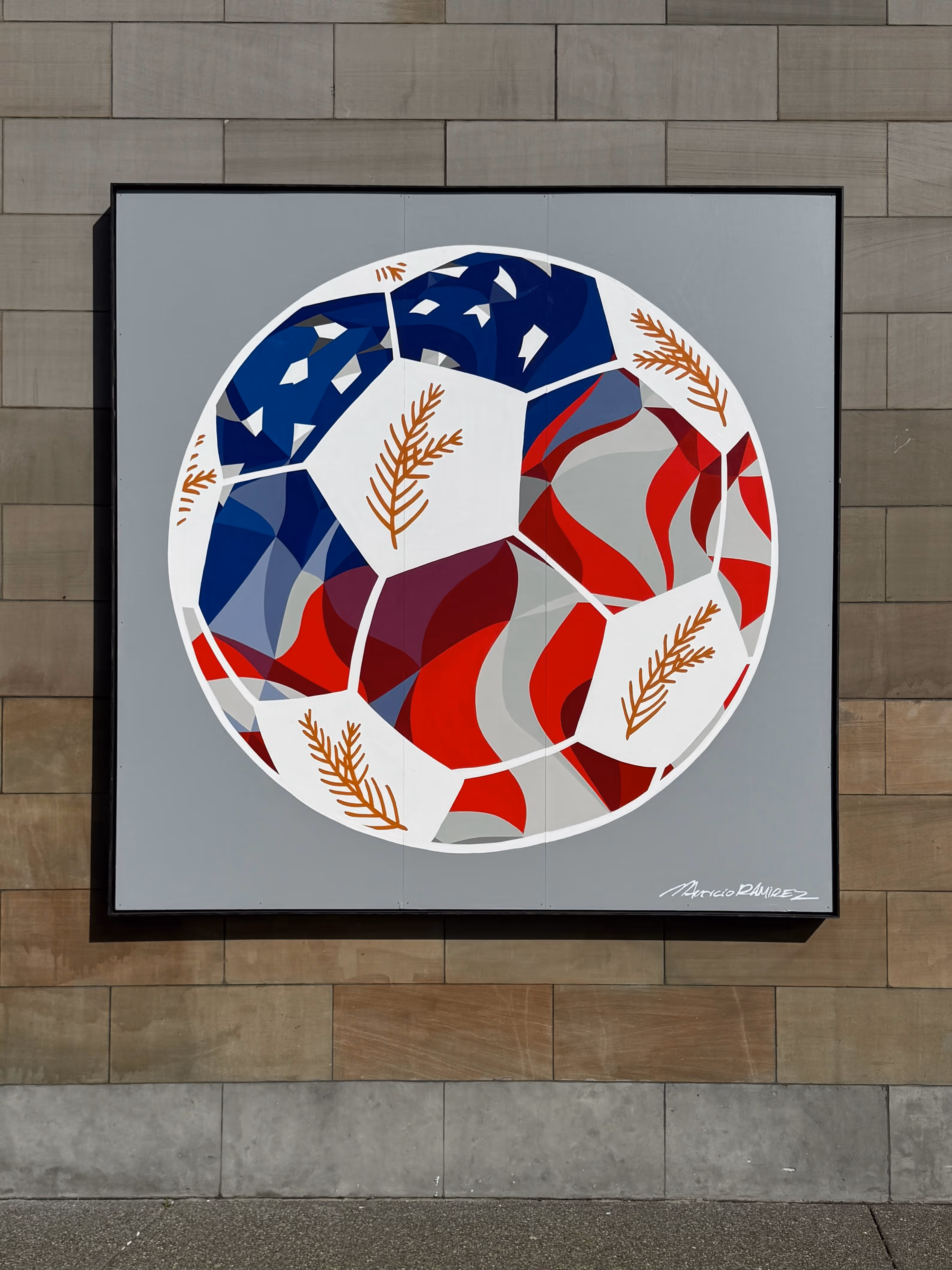 Mural of a soccer ball painted with red, white, and blue patterns and brown leaf designs on a gray background.