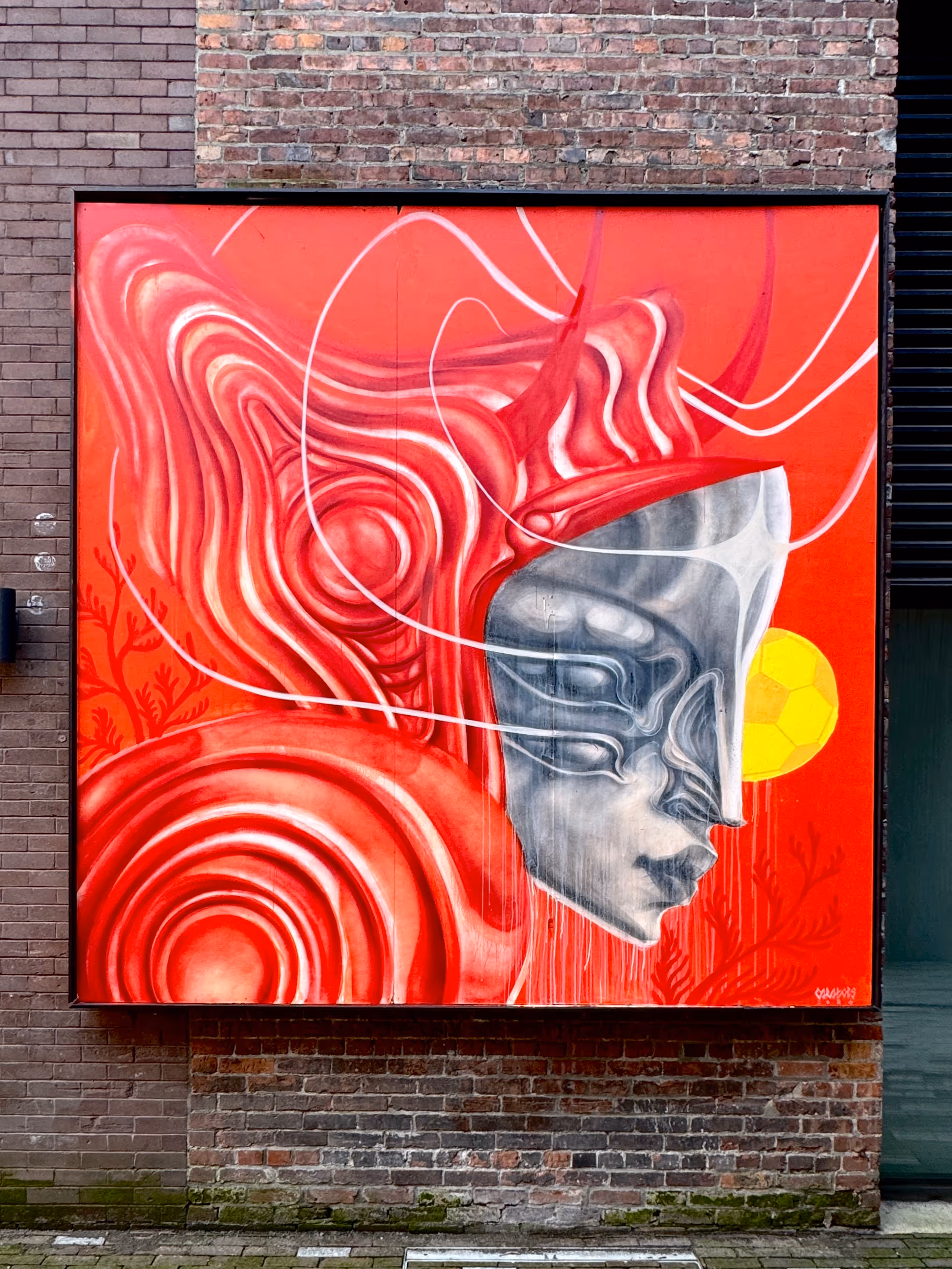 Street art of a metallic robotic face with red swirls on a brick wall background.