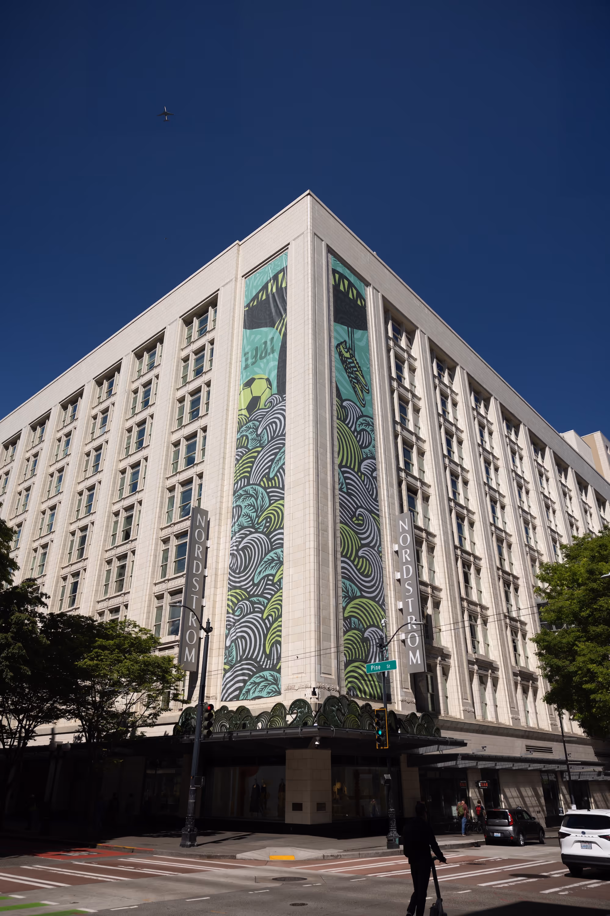 Nordstrom building corner with large colorful mural under clear blue sky and an airplane overhead.