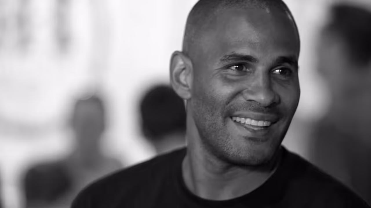 Smiling man with close-cropped hair wearing a dark shirt in a softly blurred background.