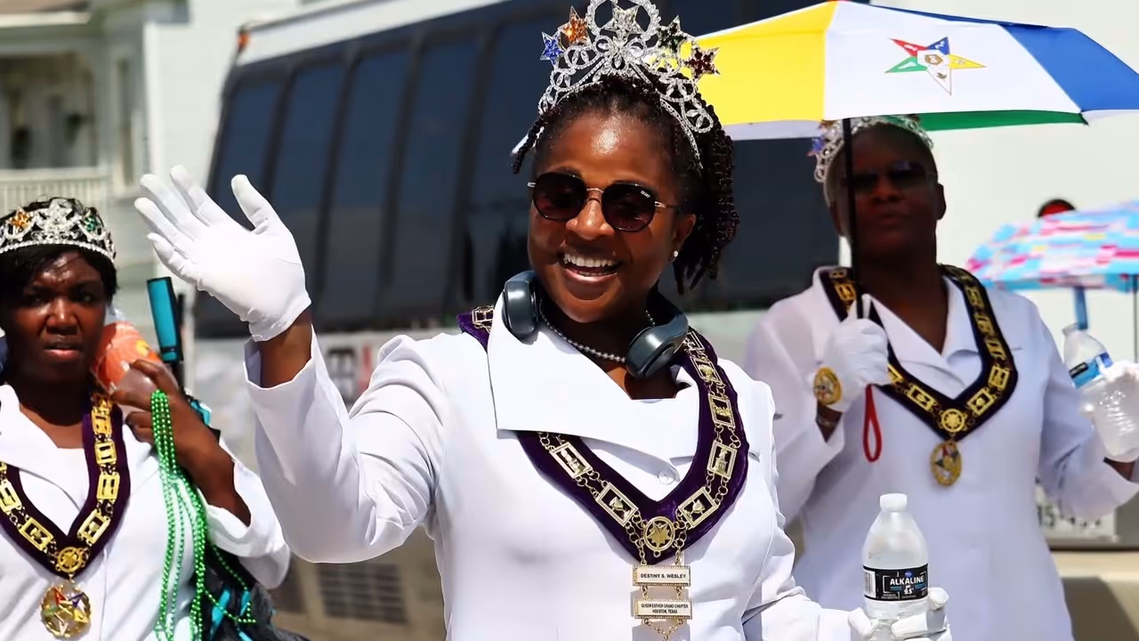 Three women in white uniforms and crowns, one waving and smiling, holding water bottles and umbrellas.
