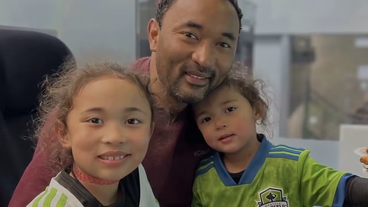 Man with two young children smiling, one wearing a green soccer jersey and the other a white one.