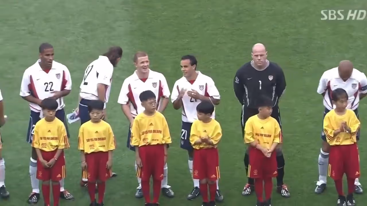 U.S. soccer players lined up on grass field with children in yellow shirts standing in front of them.