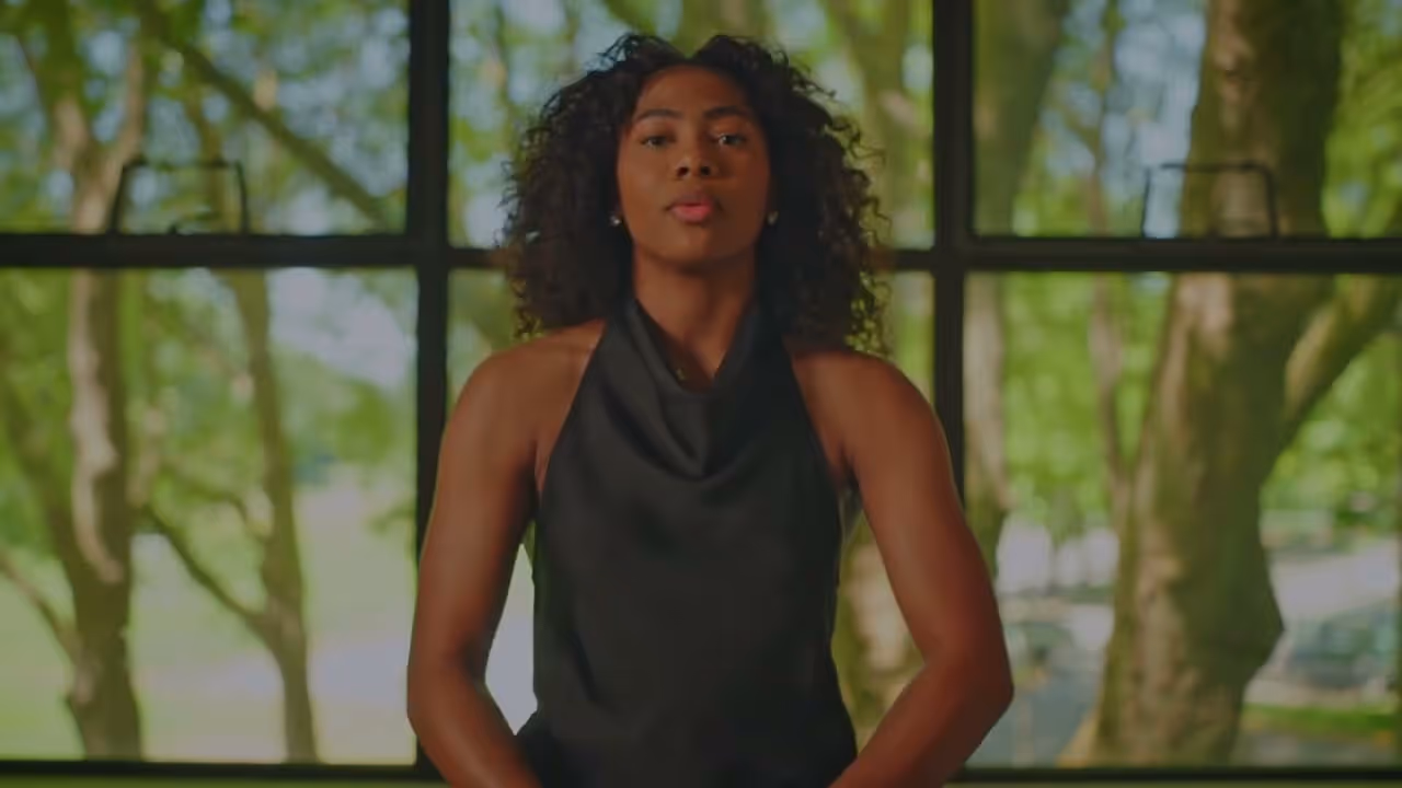 Woman with curly hair in a black sleeveless top standing in front of a window with green trees outside.