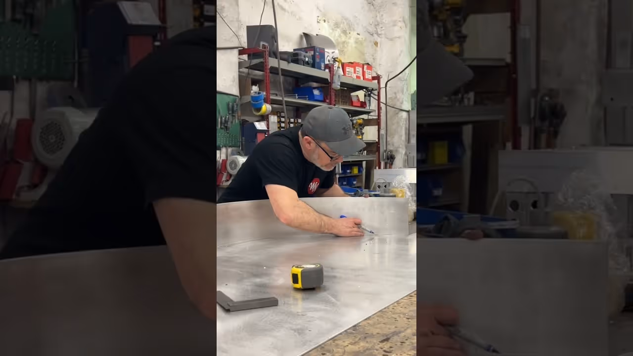 Man in a workshop marking measurements on a large metal sheet with tools around him.