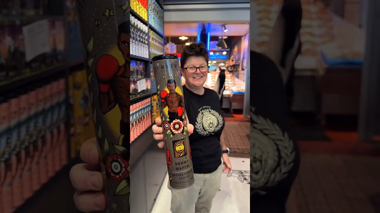 Person smiling and holding a decorated tube labeled 'Benne Wafers' inside a store aisle.
