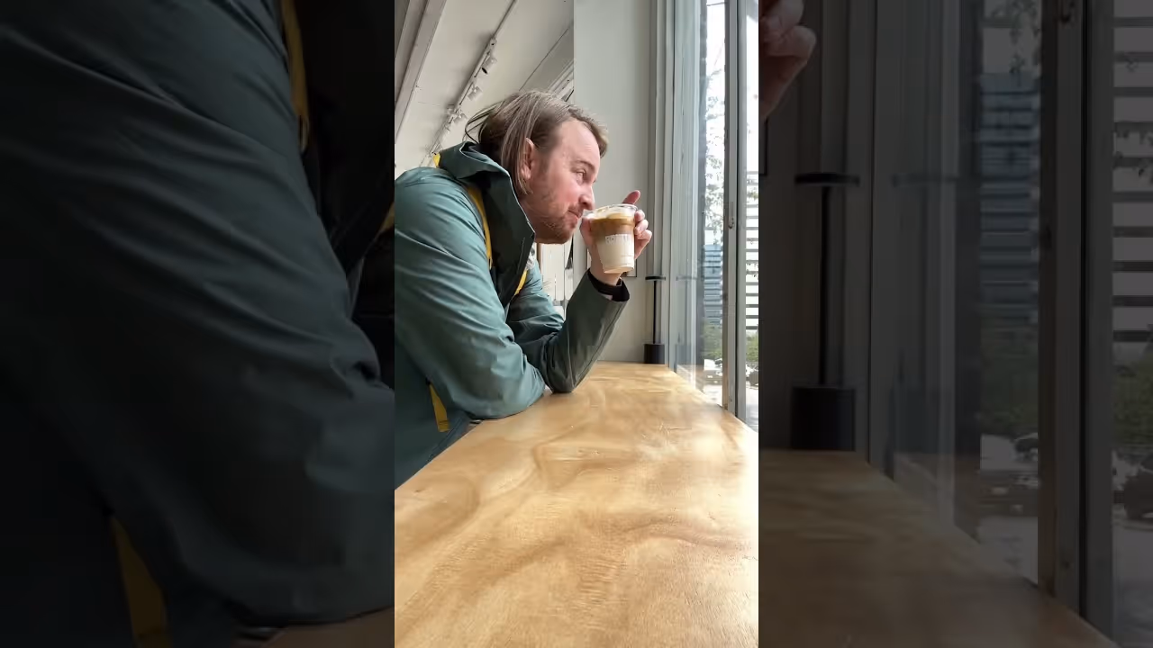 Man in green jacket drinking iced coffee by a large window at a wooden counter.