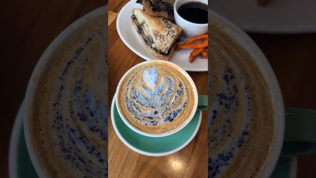 Cup of latte with floral latte art on a green saucer, with sandwich and sweet potato fries on a plate behind it.