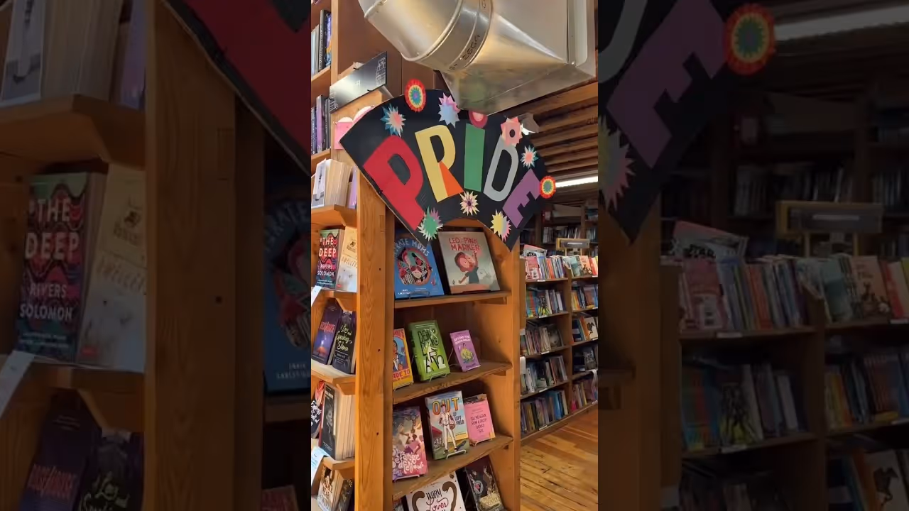 Wooden bookshelf in a bookstore with colorful PRIDE sign and various displayed books.