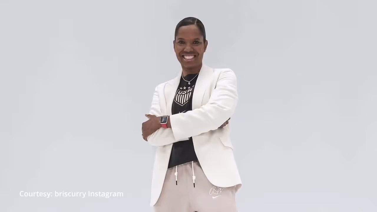 Smiling woman in white blazer and USA sportswear stands with arms crossed on gray background.