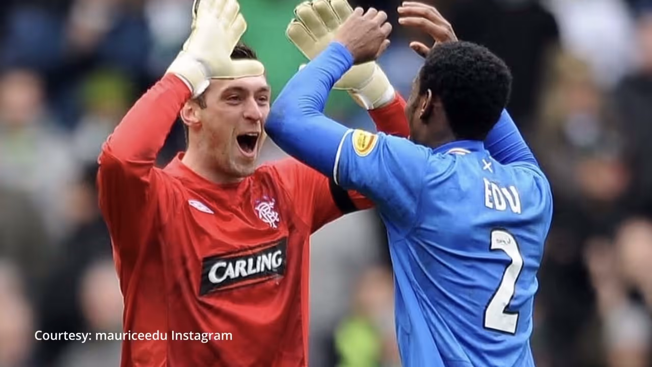 Two soccer players high-fiving; one in a red goalkeeper jersey, the other in a blue jersey with 'EDU' and number 2.