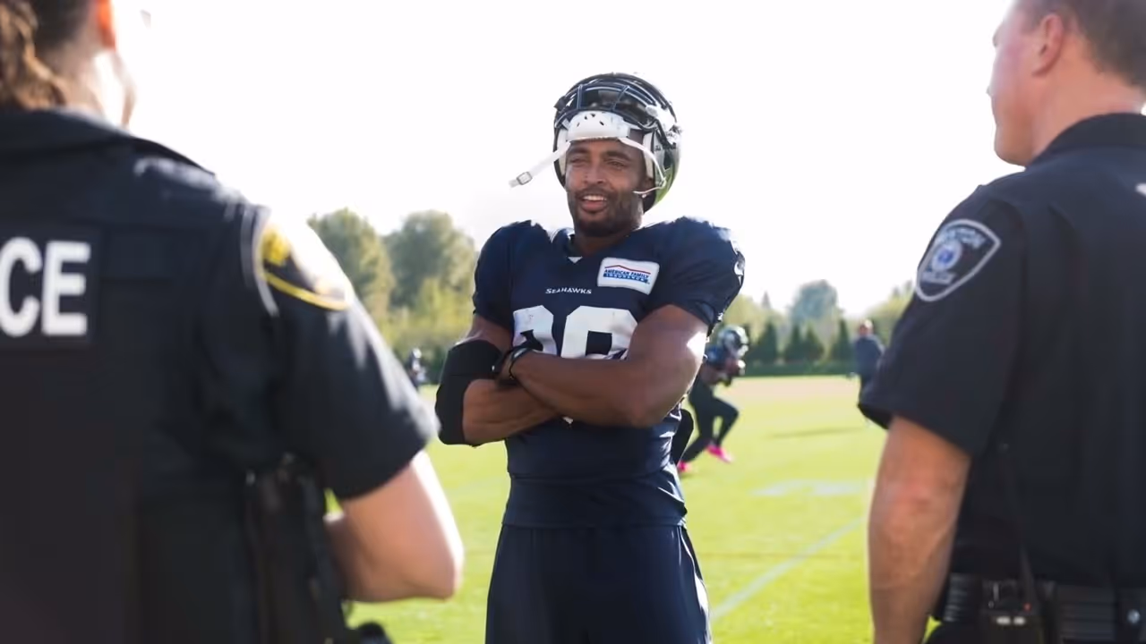 Football player in Seahawks uniform talking with two police officers on a sunny field.