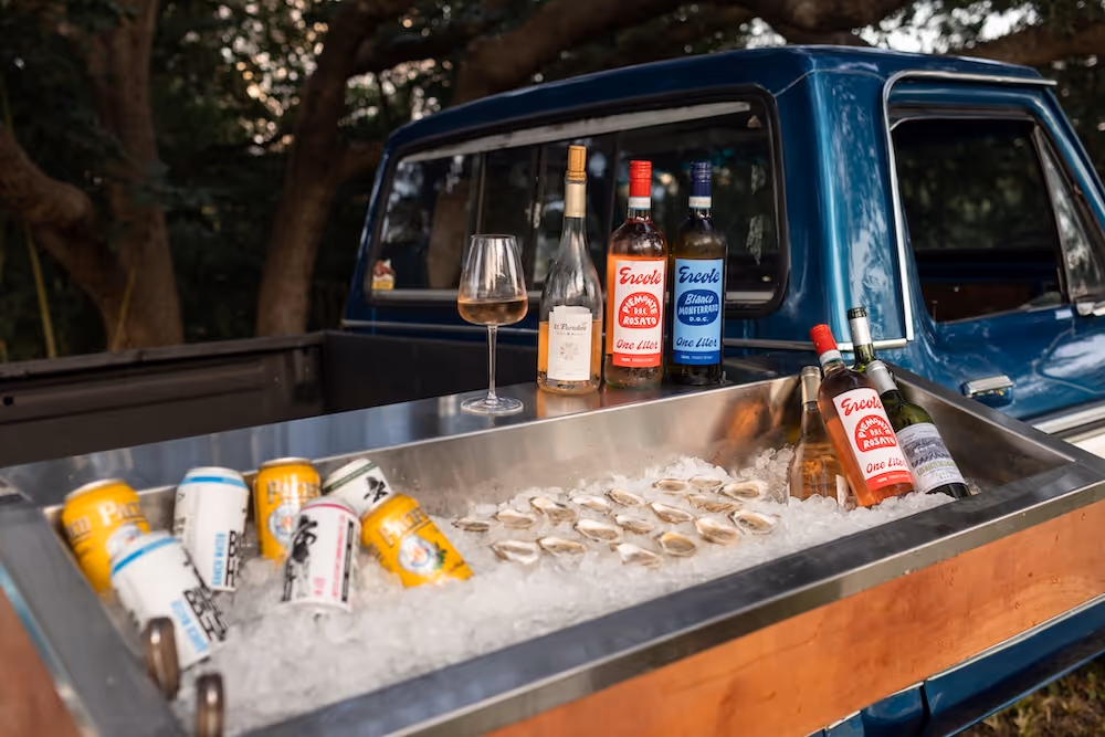 Blue pickup truck bed fitted with a wooden and metal cooler holding ice, canned drinks, wine bottles, a glass of rosé wine, and fresh oysters on the half shell.