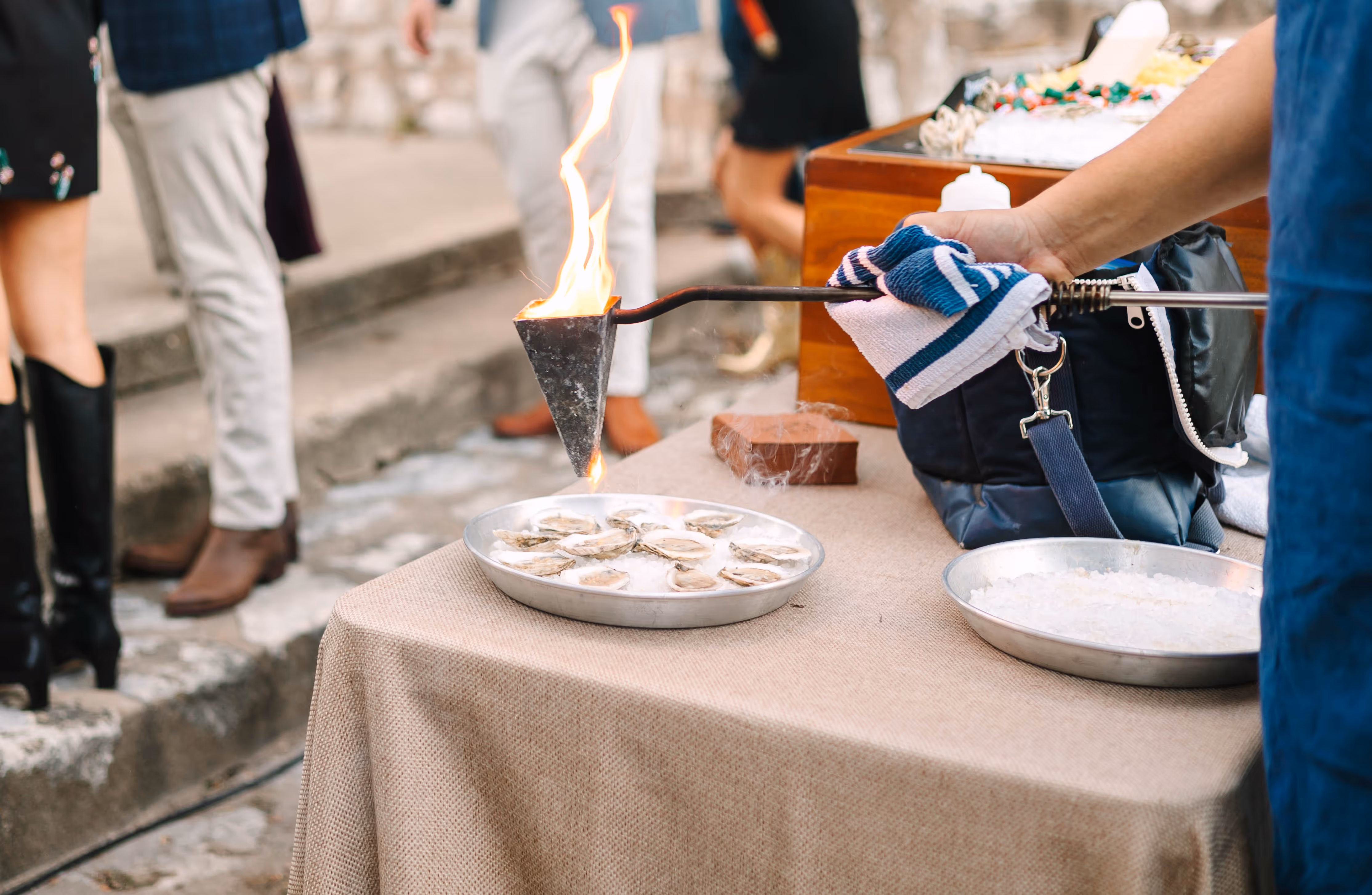 Person using a flambadou to dress oysters on a metal tray filled with ice at an outdoor event.