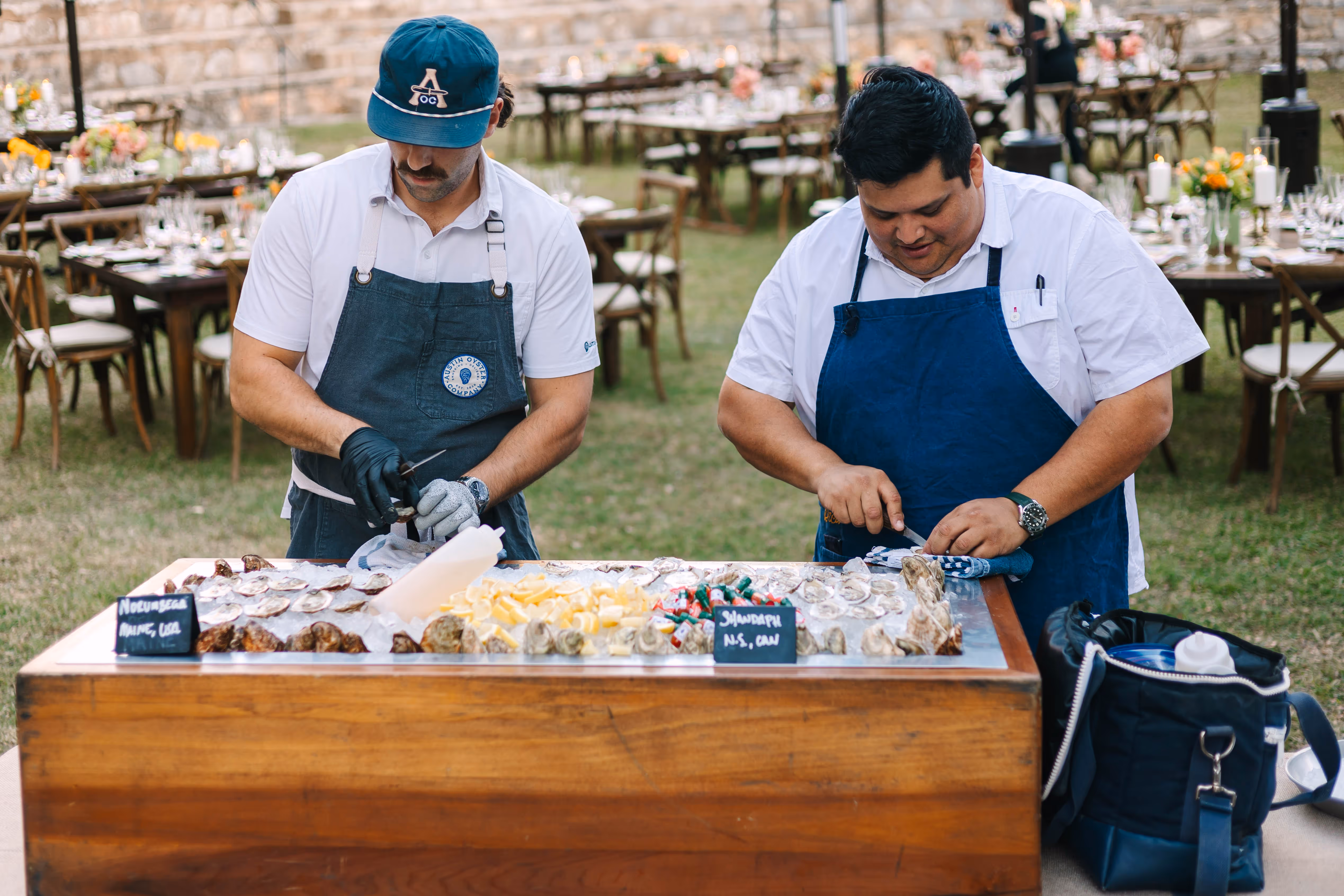 Two men shucking oysters at an outdoor event with tables set for dining in the background.