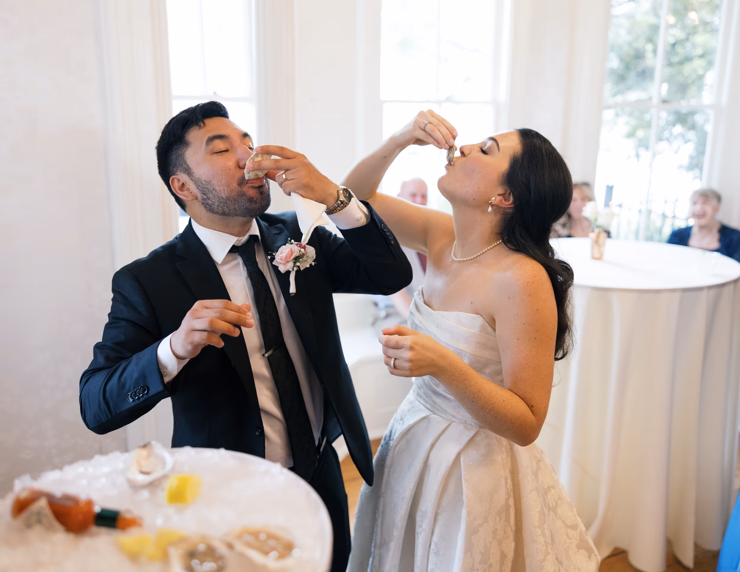 Bride in a white wedding dress and groom in a dark suit eating oysters together indoors at a wedding reception.