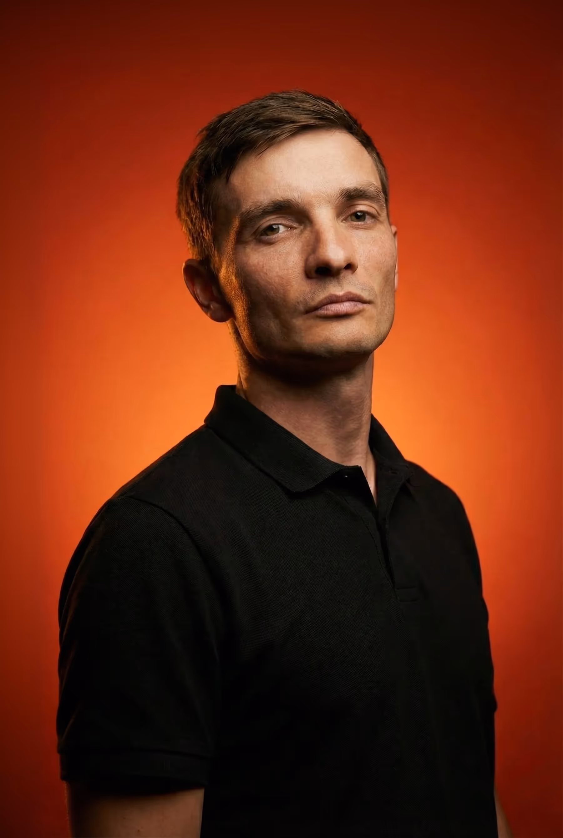 Portrait of a man with short brown hair wearing a black polo shirt against a warm orange background.