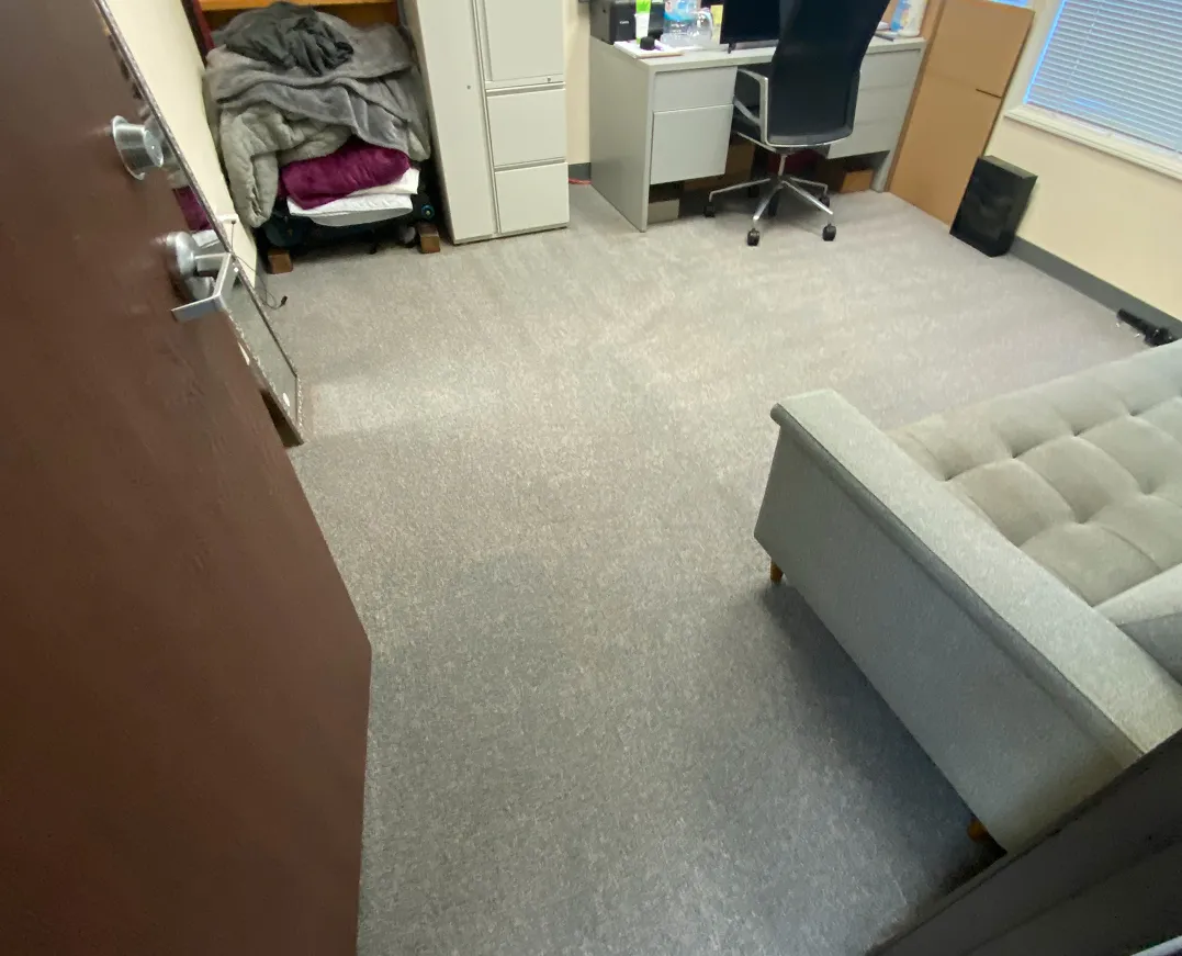 Office room with grey carpet, grey tufted sofa, desk with black office chair, and stacked blankets on a cart.