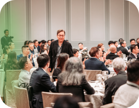 Person walking between tables in a conference room filled with attendees in business attire clapping.