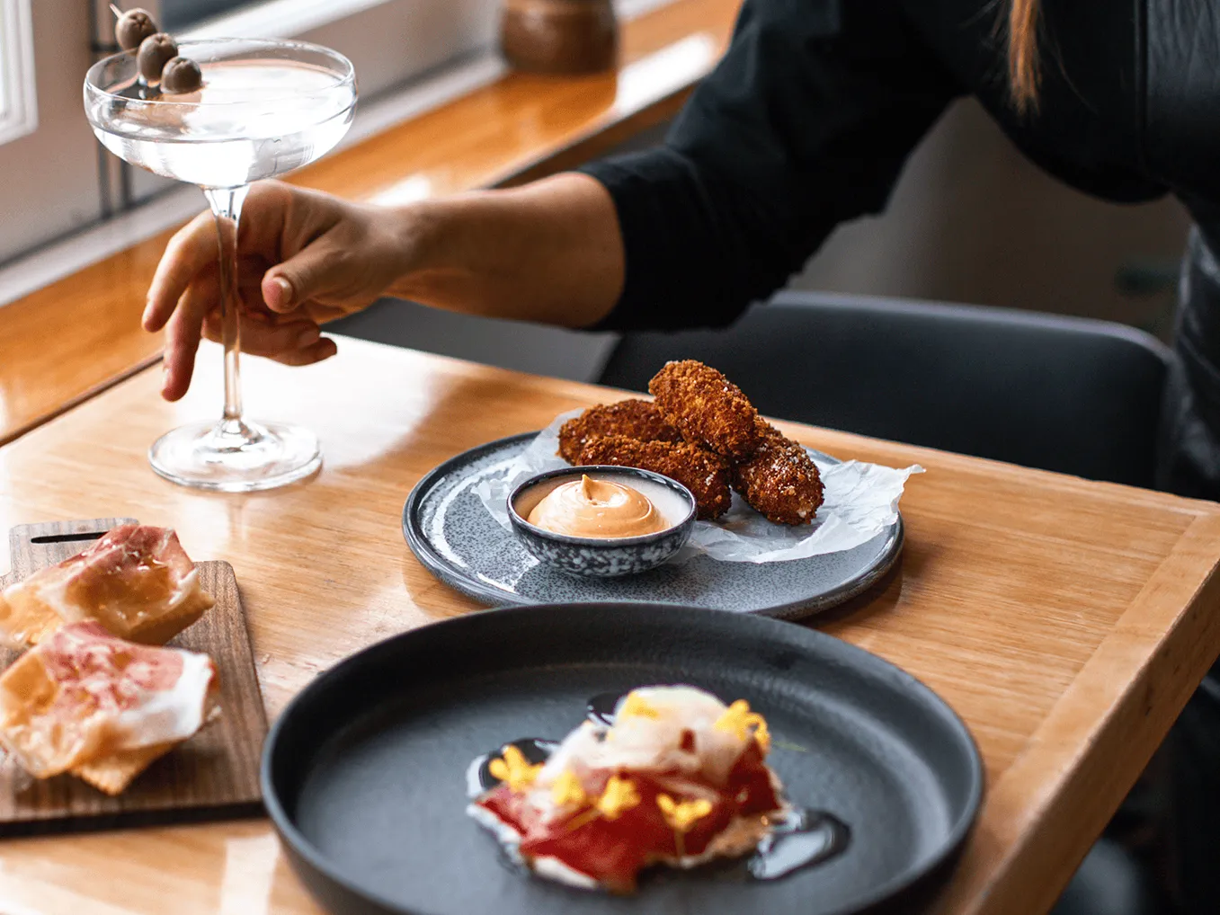 A hand holds a martini over a table full of food infront of a window.