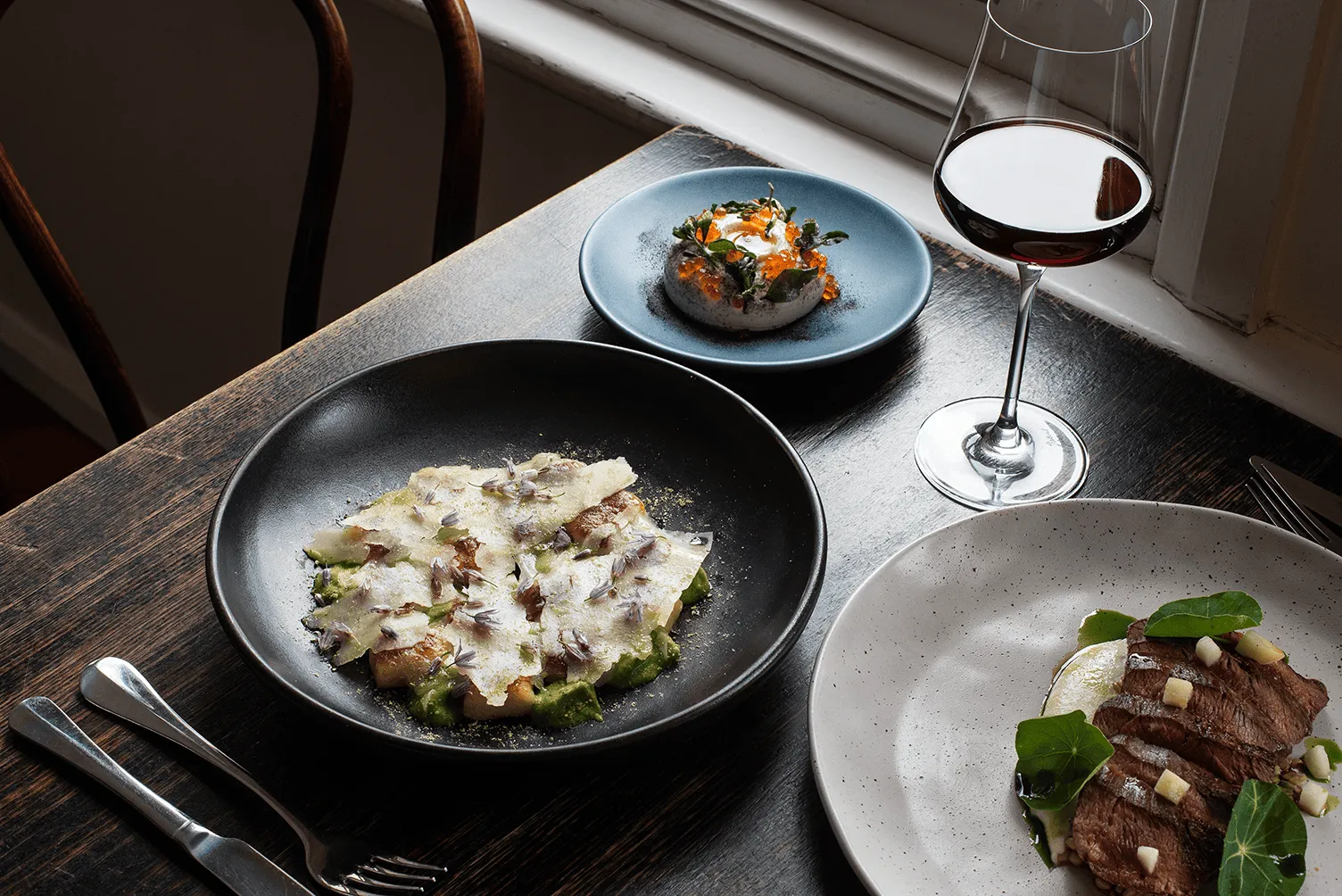 A wooden table with an assortent of delicious food. A glass of red wine. A mouse with herbs and salmon roe, a rare steak carved over mash with green nasturtium leaves. Shaved parmesan and flowers. 