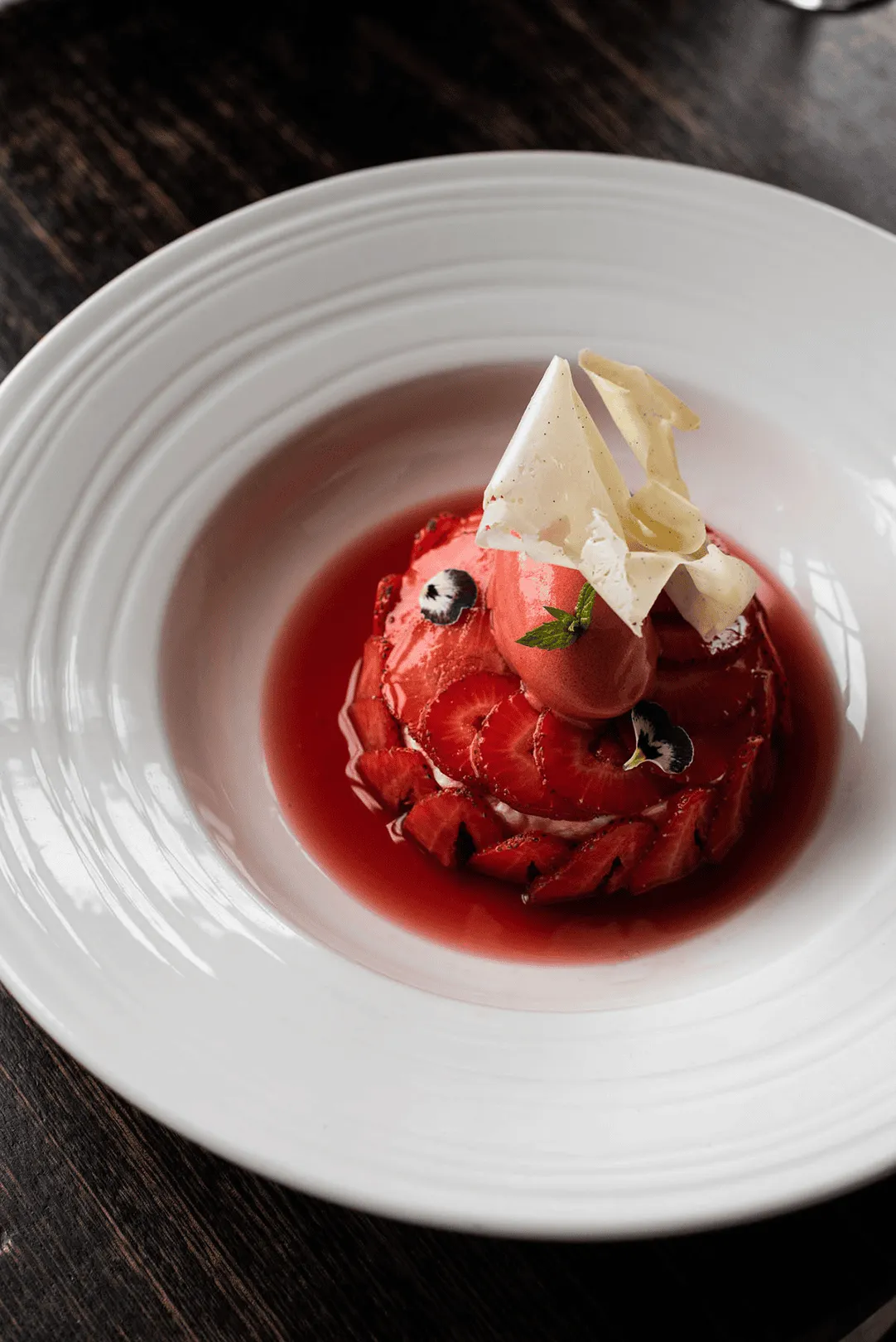 White plate, beautiful red dessert featuring strawberry fresh fruit and sauce and sorbet, garnished with flower petals.