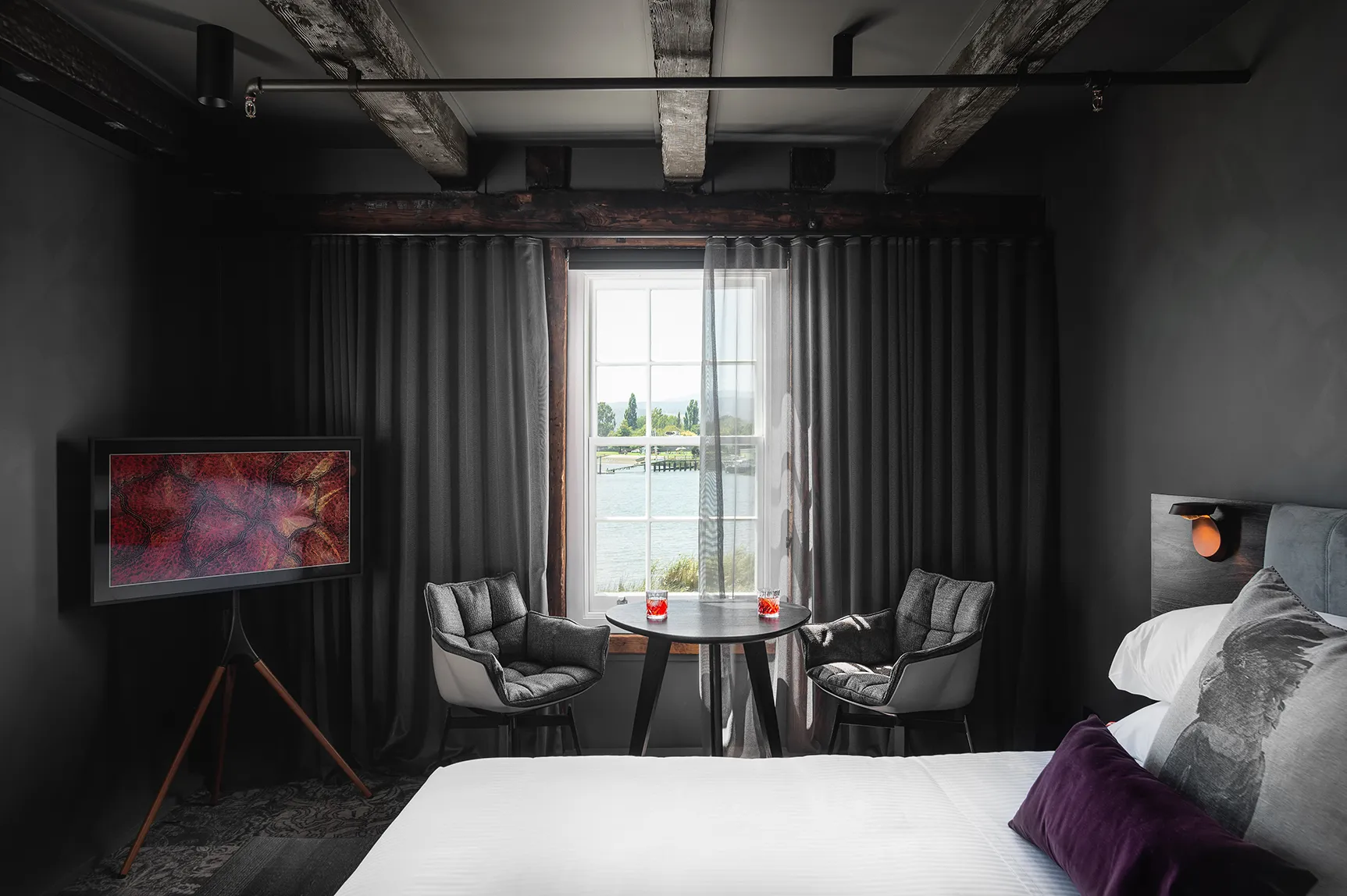 A hotel suite, with comfy bed and elegant TV. There are draped curtains infront of a windo overlooking the rivers edge. There are two chairs and a table with two negroni cocktails. Wooden beams feature on the grey walls. 