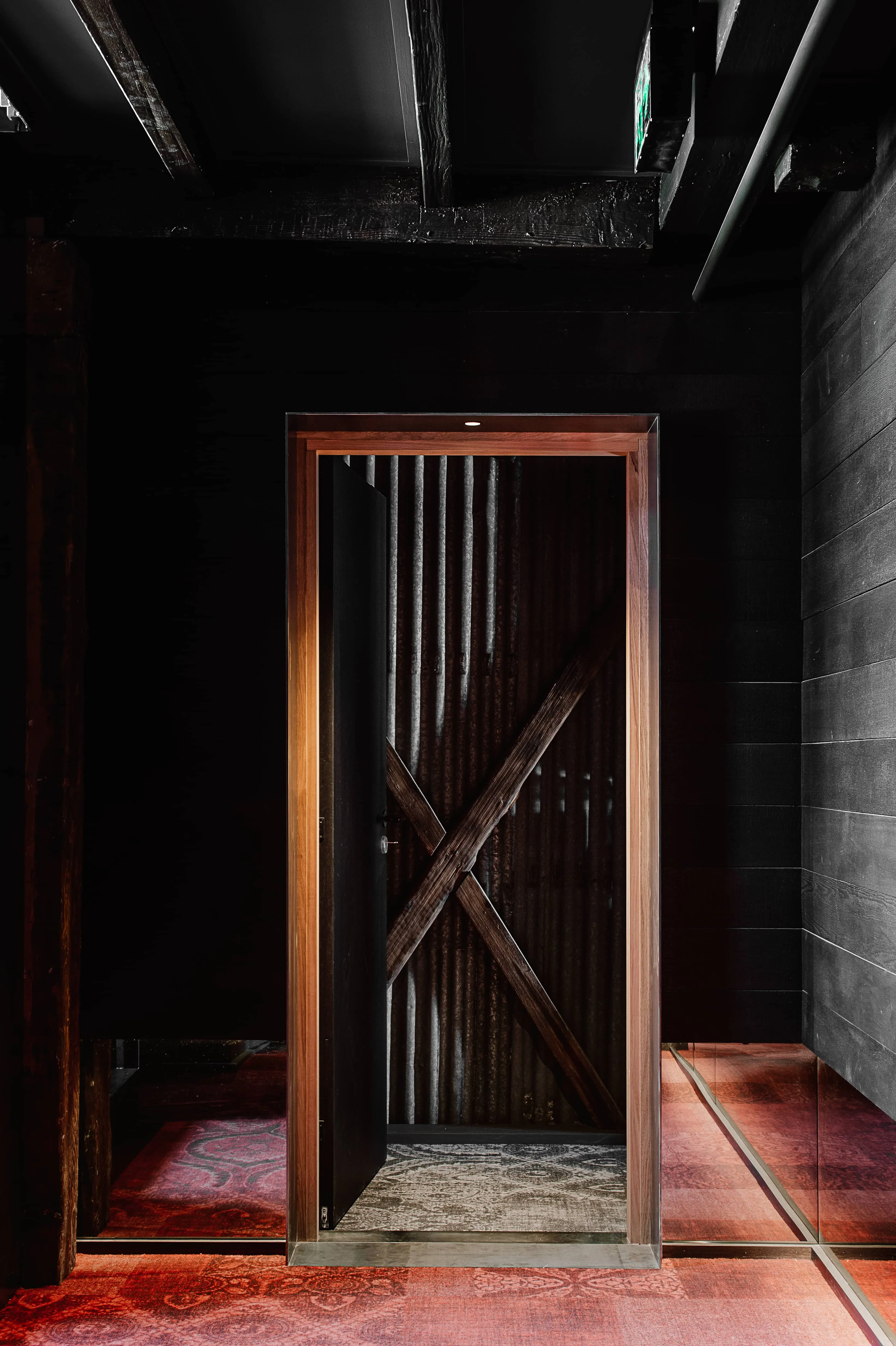 A partially open door in a dimly lit room reveals a rustic interior with corrugated metal and wooden beams. The floor is patterned with red and brown hues, and a ceiling exit sign is visible.