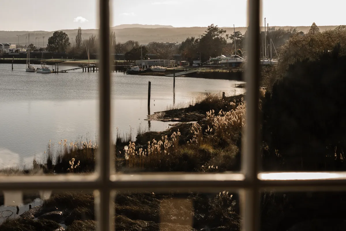 A view through the wooden window, overlooking the Tamar river shoreline with boats and hills in the background. 