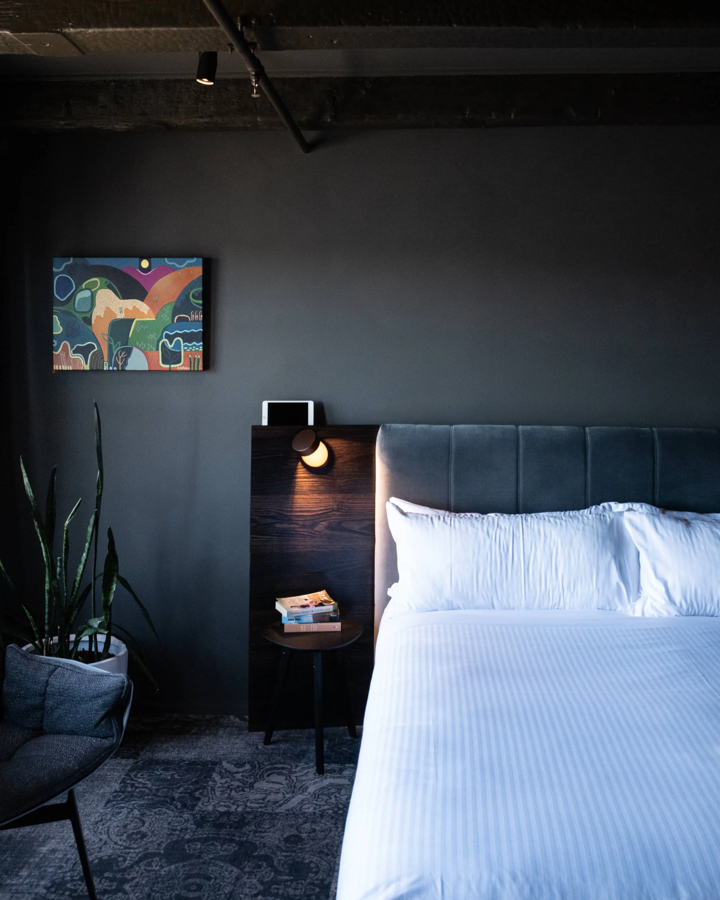 A cozy bedroom featuring a neatly made bed with decorative pillows, a bedside table with a lamp, and various bedding elements. The room has warm lighting, simple interior design, and is elegantly arranged for comfort.