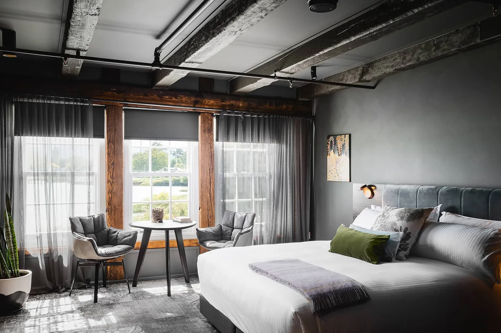A cozy bedroom featuring a neatly made bed with decorative pillows, a pair of stylish chairs positioned nearby, soft linens draped over the mattress, and elegant curtains framing a window that allows natural light to fill the space. The room has an inviting atmosphere with thoughtfully arranged furniture and tasteful decor.