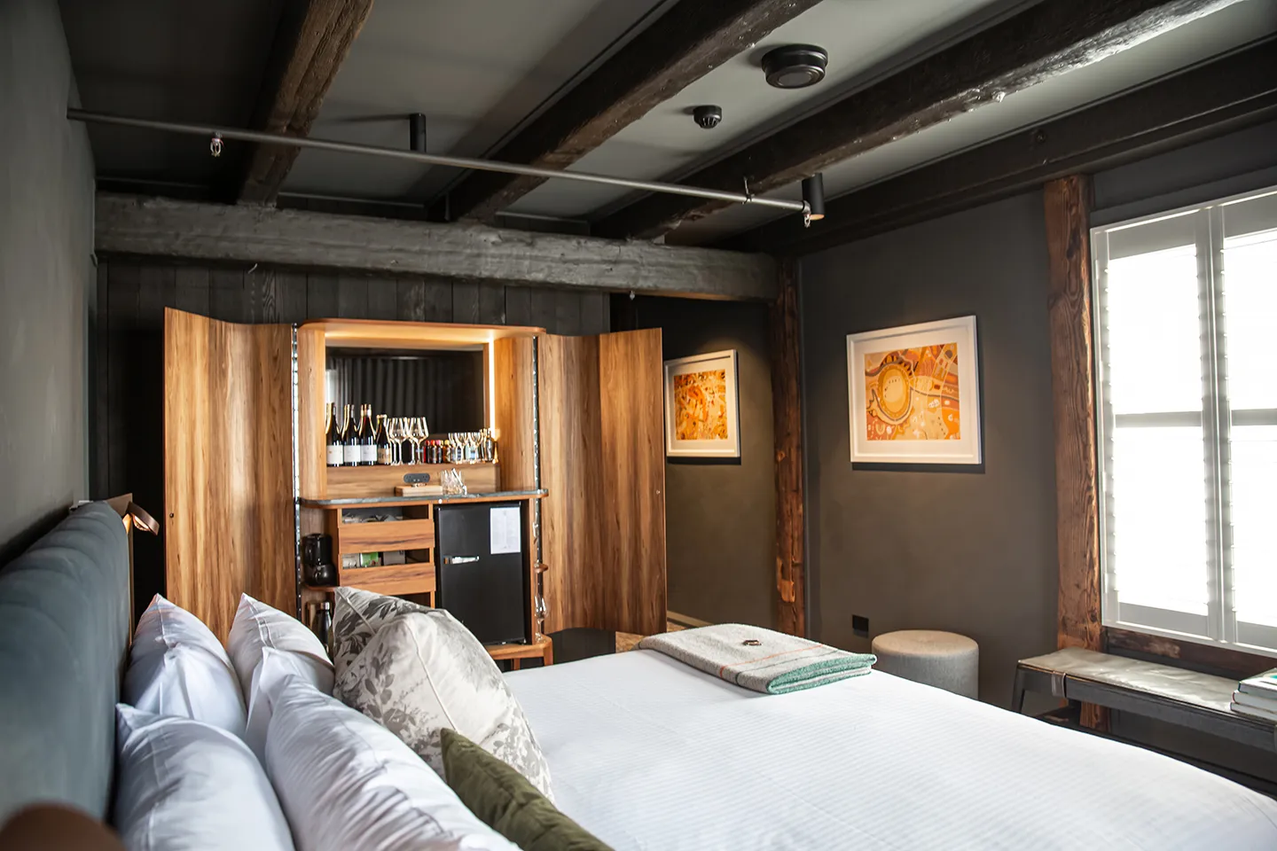 A room with a comfy bed and yellow artwork. Wooden beams show in the ceiling. An open, crafter wooden cabinet showcases a range of beverages. 