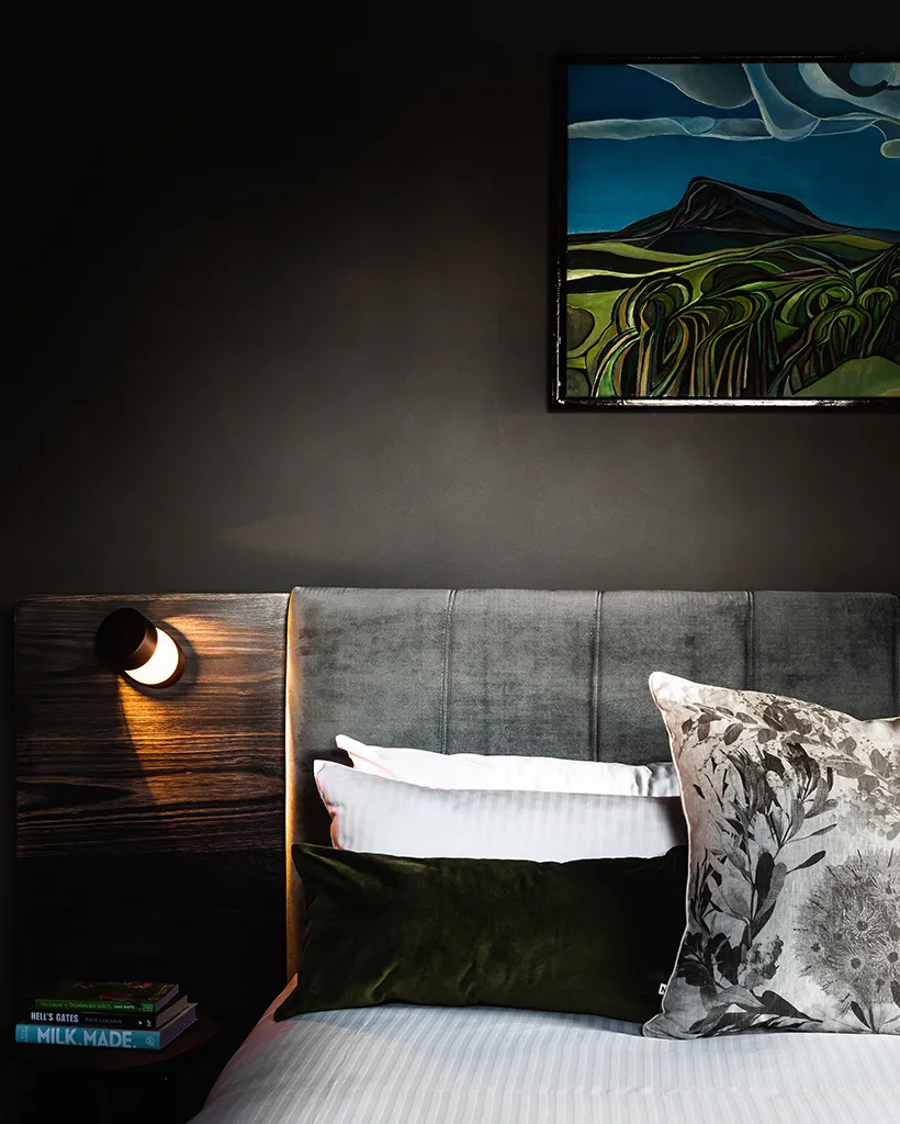 A cozy bedroom featuring a bed adorned with pillows, alongside a wall displaying an artistic painting. A cat is comfortably resting on the bed, adding to the warm indoor atmosphere.