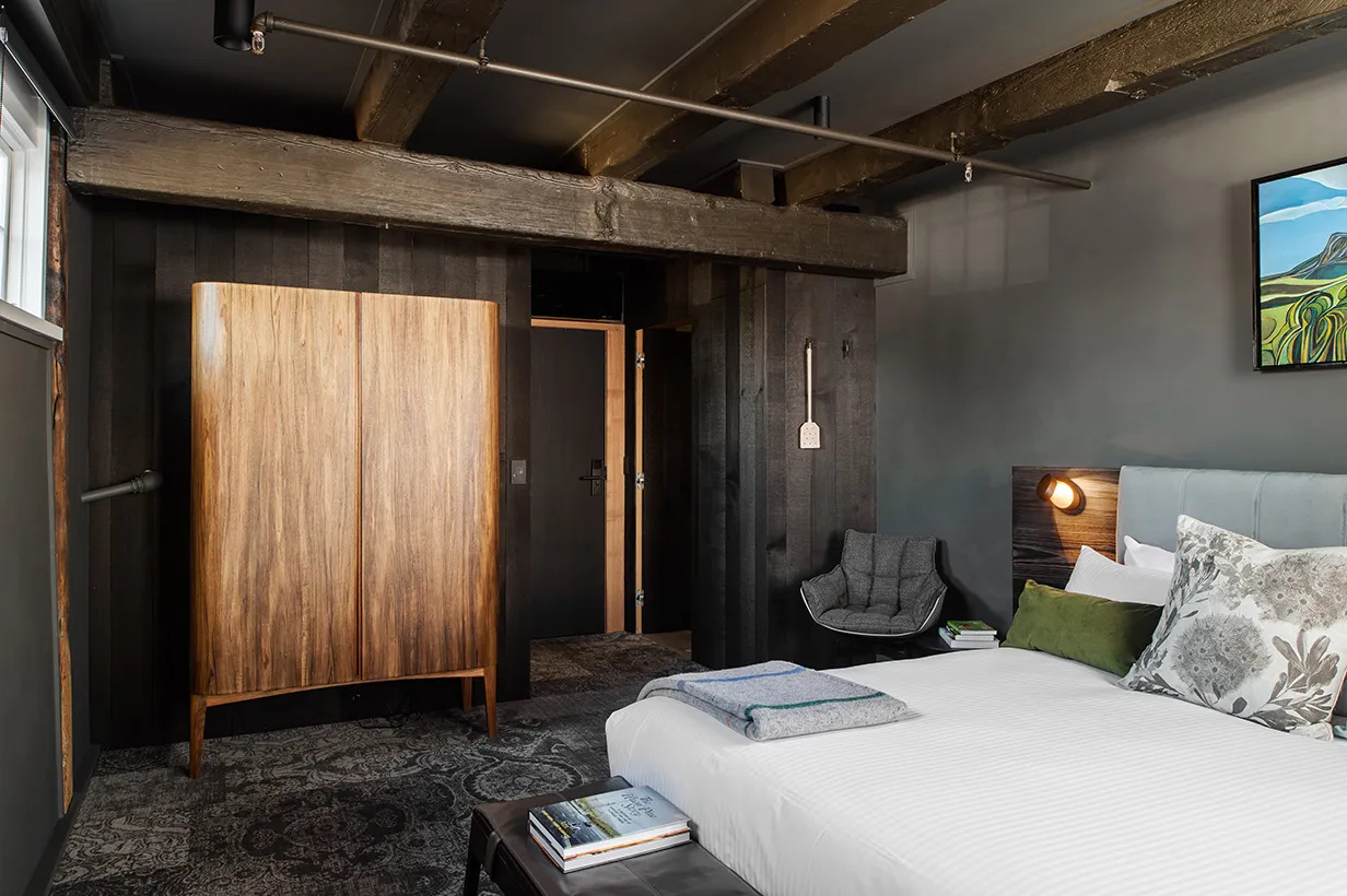 A cost room. A bed in the foreground, with green velvet cushion, warm bedlight, and a cosy chair. The room is grey tones, with a patterned grey carpet and a wooden crafter drinks cabinet. Wooden beams can be seen, a can the door to the room. There is a low table with a book on it, and a warm woolen rug. 
