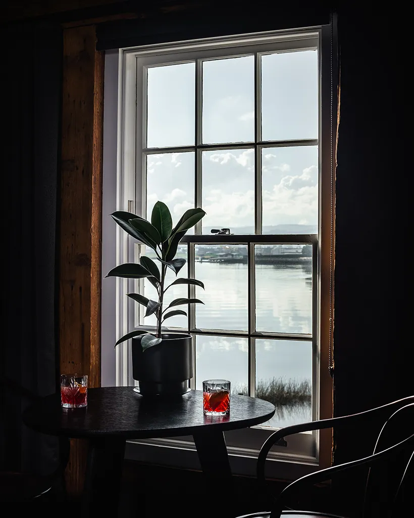 A bentwood chair sits in front of a window. On the table is two negroni cocktails and a plant. The view through the wooden, grided window frame is of the river, showing relfections, boats and reeds. 