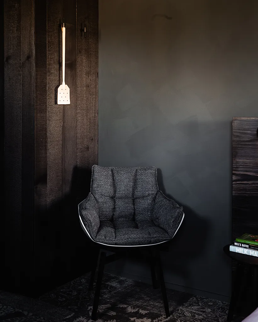A comfy chair sits infront of a grey wall. Timber features with a leather fly swat can be seen. As can the side of a black wooden bedframe, and a stack of books.