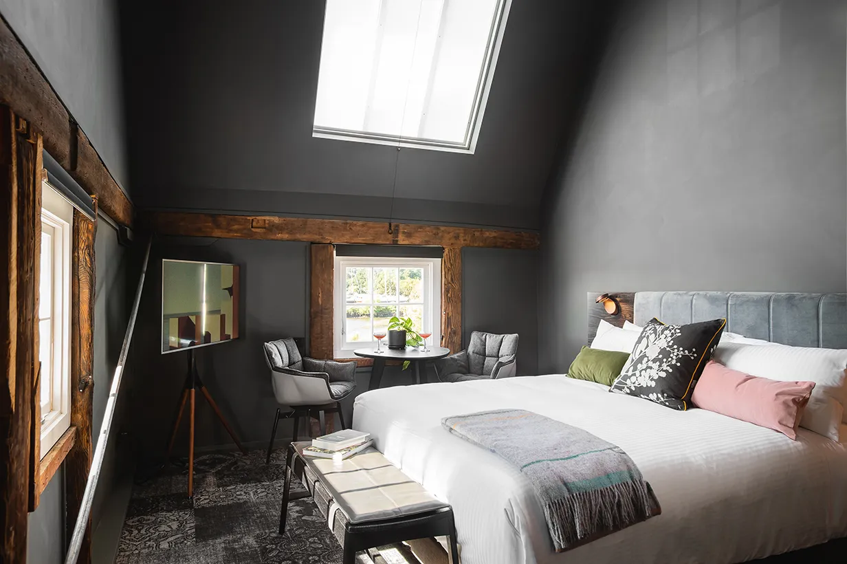 A cozy bedroom featuring a bed with pillows and a duvet cover, a desk positioned beside it, and natural light streaming in through a window. The room is decorated with a nightstand and lampshade, showcasing an inviting indoor space suitable for relaxation.