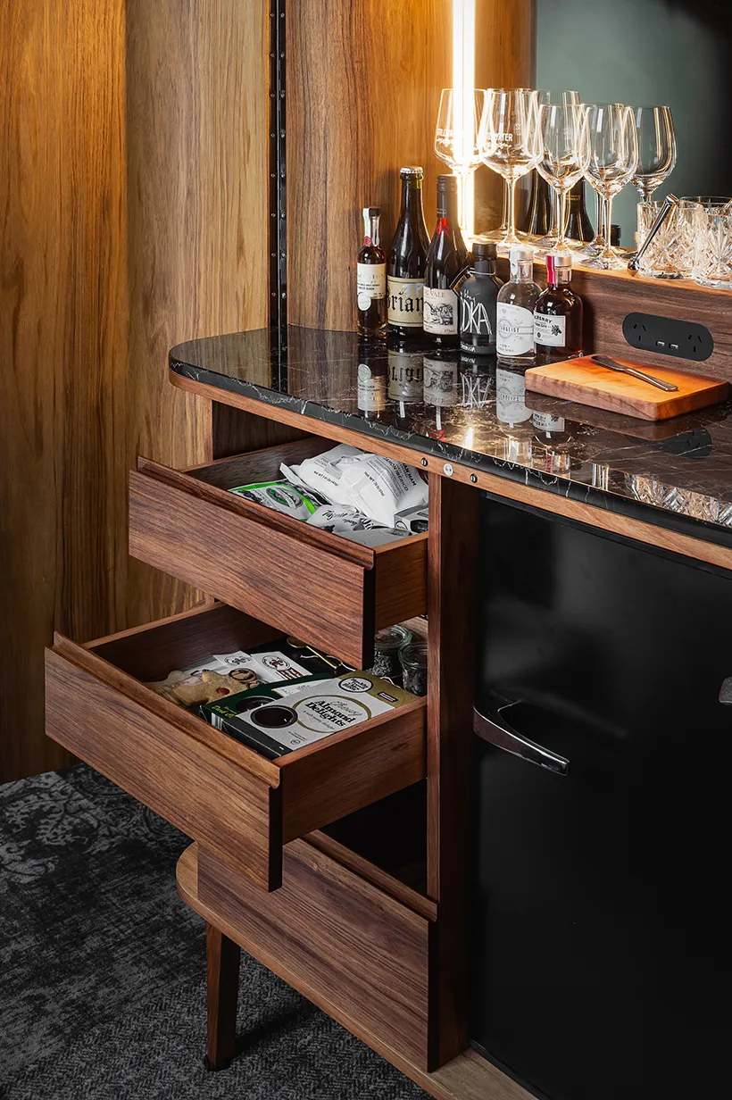 A beautifully crafted wooden drinks cabinet has its draws open. There a many delicious things in the draws. And a range of tasting wines, spirits and a row of wine glasses ontop of its marble top. 