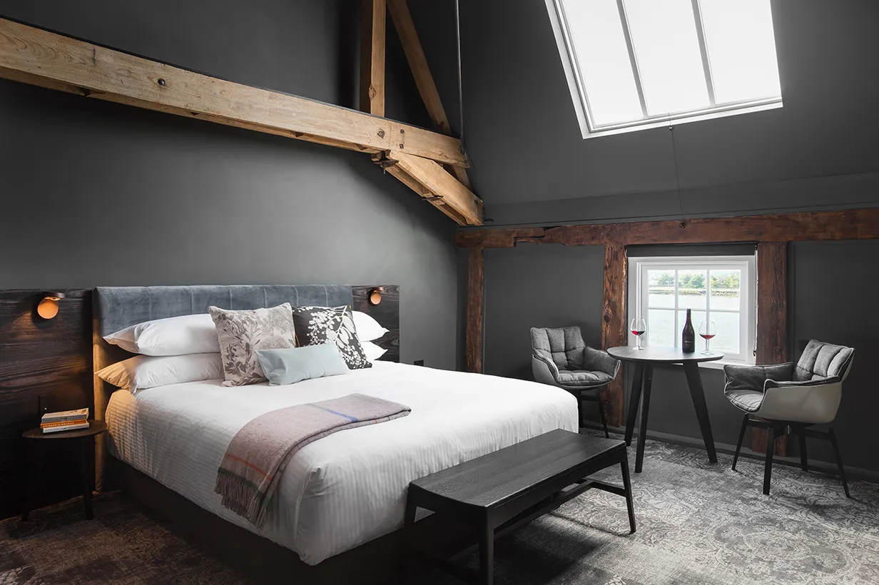 A cozy bedroom featuring a bed with plush bedding, decorative pillows, and a nightstand beside it. There are two chairs situated in the room, surrounded by hardwood flooring and a soft atmosphere enhanced by natural light coming through the window. The walls are elegantly decorated, complementing the overall interior design of this intimate space.