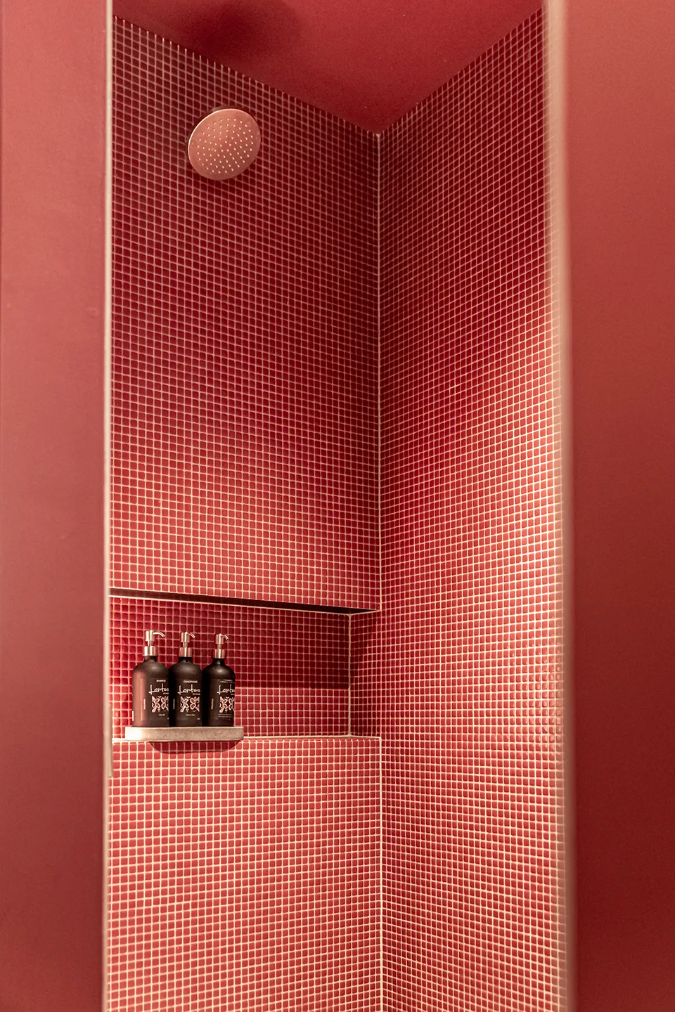 A shelf mounted on a wall, displaying several bottles of various shapes and sizes. The background features a bathroom setting with tile walls, a mirror, and a shower door. A shower room with pink ceramic tiles. 