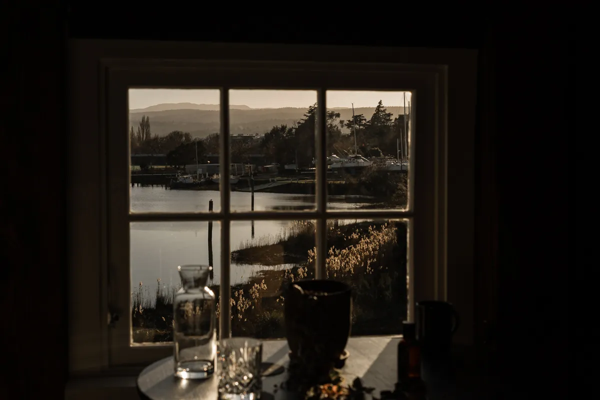 A view through a window overlooking the rivers edge. Reeds and boats can be seen through the gridded wooden window panels. A table with glasses is blurry in the foreground. 