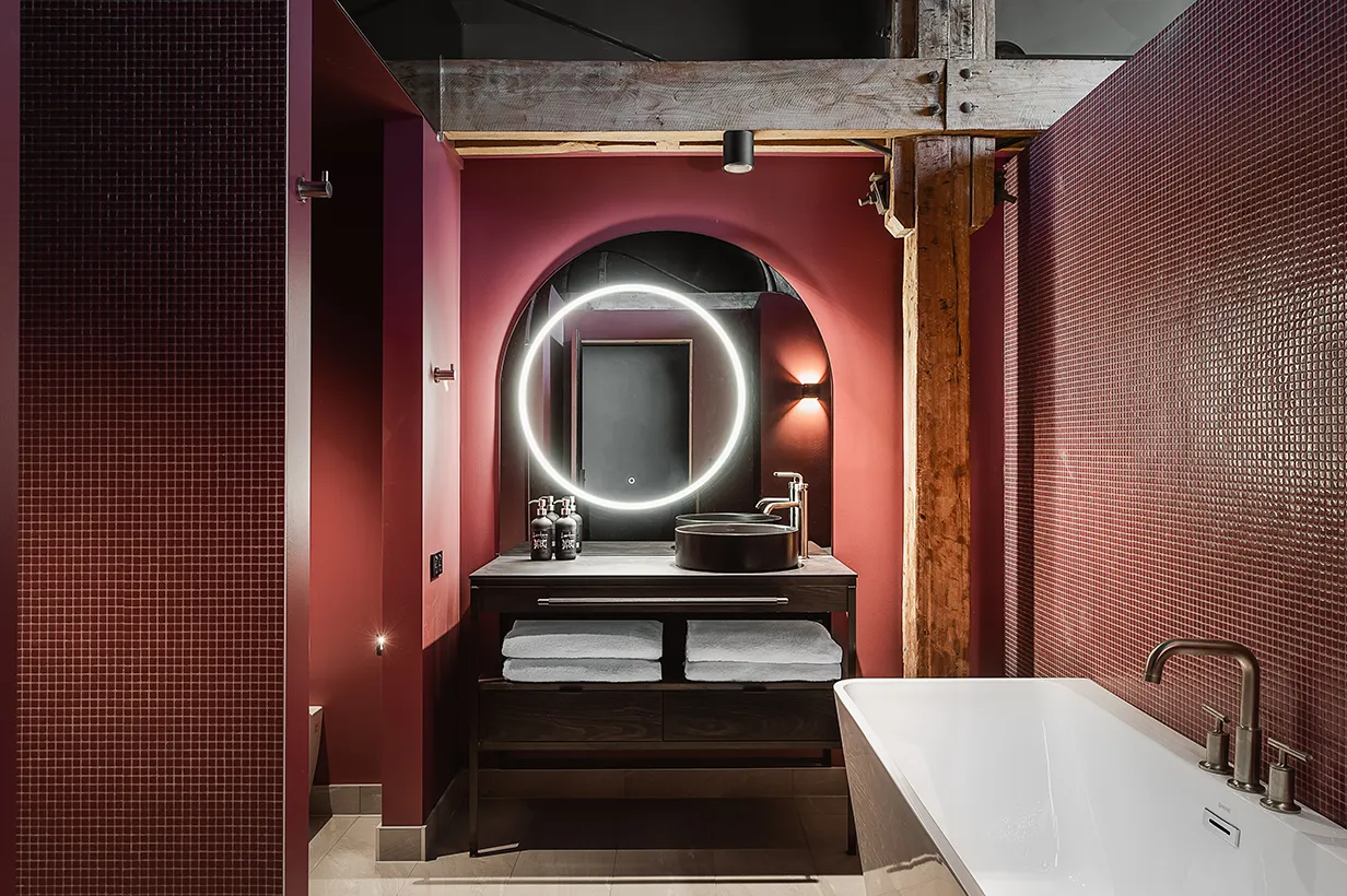 A beautifully designed bathroom. Pink tiles and a circular light infront of a large mirror. A bath is in view, as is the wooden beams, and a circular basin on top of a cabinet. 