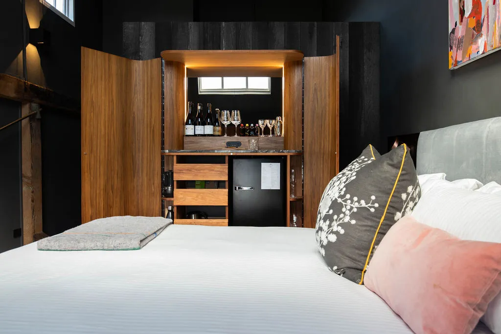 A comfy bed sits in the foreground of a hotel room. A beautiful crafter wooden drinks cabinet is open, shoping wine, glasses and cocktails. Colourful art is on the wall. 