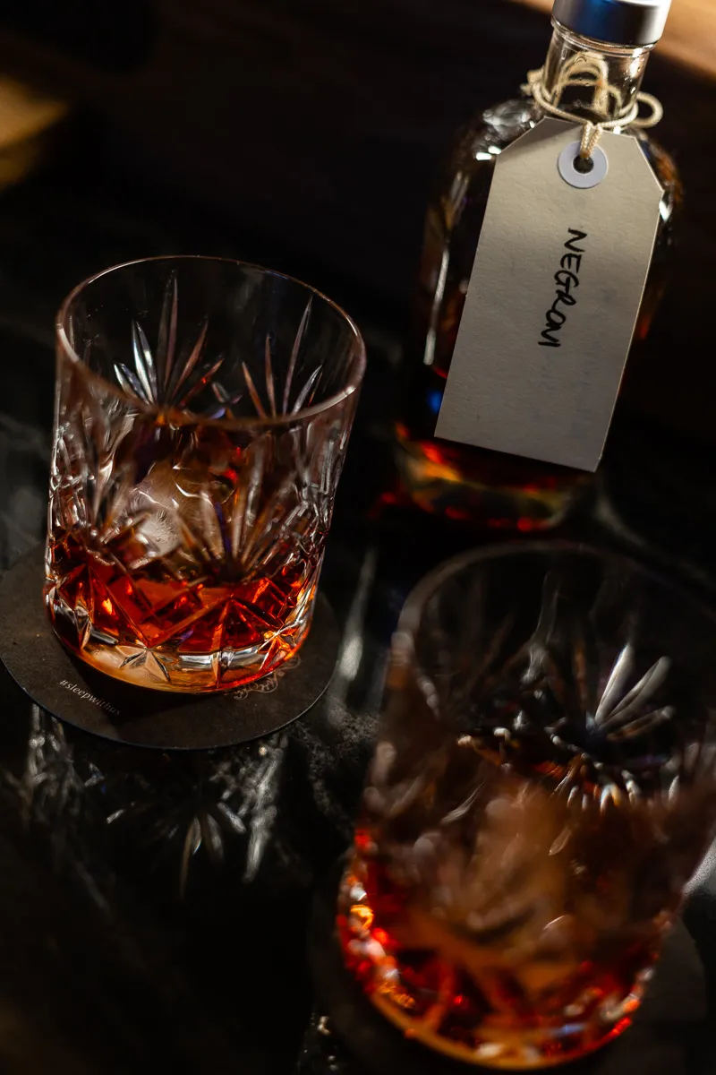 Closeup of two glasses of negroni cocktail in a faceted glass. A small cocktail bottle sits behind with the word Negroni written in black handwriting on a label. 